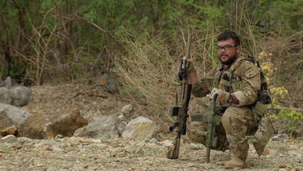 Obraz premium A special forces soldier kneels in a rocky wilderness, holding two assault rifles while executing a tactical reconnaissance mission for combat readiness.