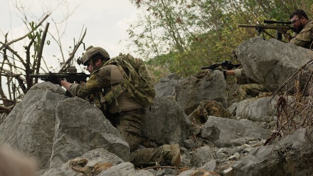 Special forces soldiers in full combat gear take defensive positions behind rocks, aiming their rifles during a tactical reconnaissance mission in hostile territory.