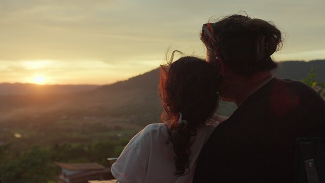 A mother and daughter watch the sunset over mountains from a viewpoint, sharing a quiet moment of family bonding and appreciating the tranquil scenic view.