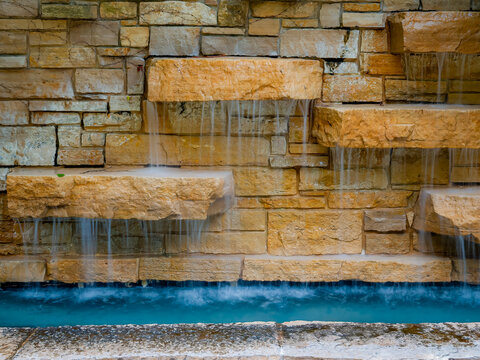 Modern outdoor stone brick wall water feature with cascading tiered waterfalls flowing into a clear blue swimming pool