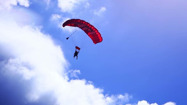 Tandem skydivers descending under red parachute against vibrant blue sky and white clouds during extreme sports jump.