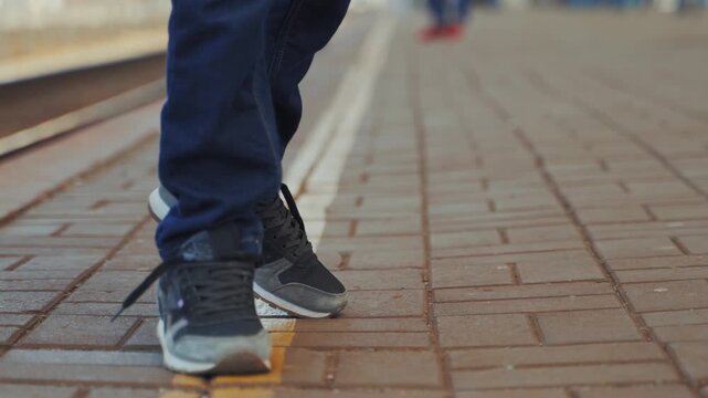 A young boy in blue jeans and black sneakers walks along yellow safety line on a train platform, with other blurred people in background.