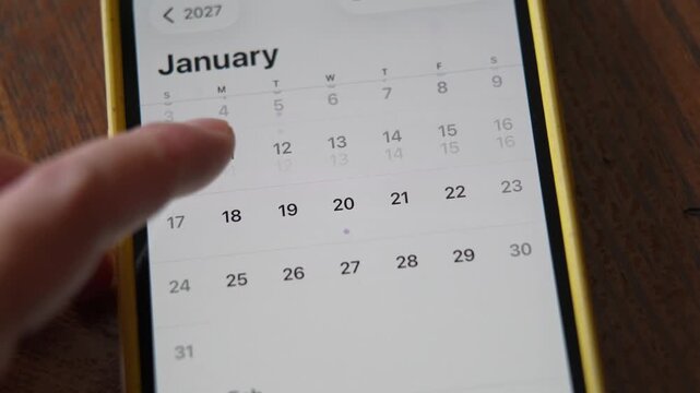 Female finger is scrolling calendar app on mobile phone screen looking for date. Close up. 2027 year. Search for Valentine's day. Electronic calendar in a smartphone.