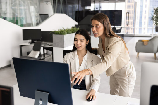 Young 30s Latin female coach or mentor provide explanations about new program or software to young Asian woman colleague, reviewing data on computer screen, pointing on details, engaged in discussion