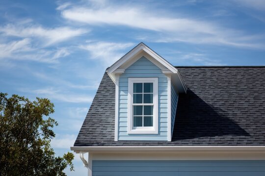 Close-up of a dormer window on a light blue residential house with a grey asphalt shingle roof against a clear blue sky.