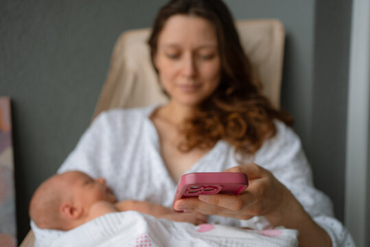 New mother balancing childcare and technology, cradling her sleeping newborn baby while engaging with a smartphone, representing modern parenting in the digital age