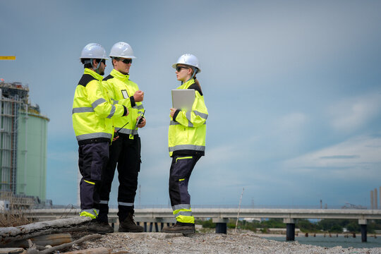 Construction worker safety hardhat high visibility jacket clipboard industrial site inspection teamwork, focused engineer group discuss safety plan near bridge and plant, outdoor supervision
