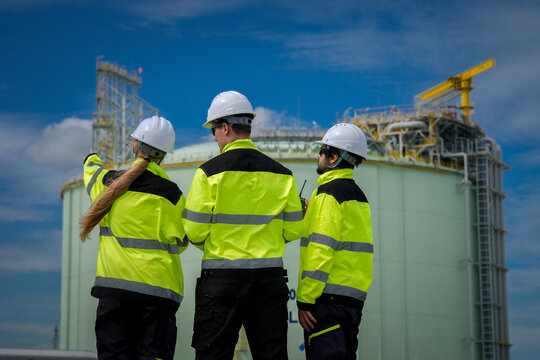 Industrial worker safety hard hat high visibility jacket inspection storage tank team at energy facility, back view, pointing at equipment, professional focus under blue sky