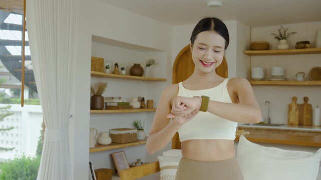 Beautiful young Asian woman in activewear smiles while checking her fitness tracker smartwatch at home. Healthy lifestyle, wellness, and modern technology for daily sports and workout routines.