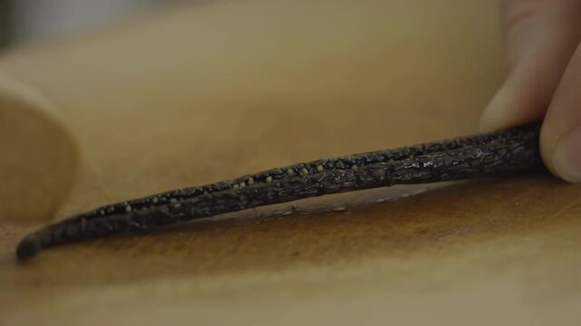 Close-up of hands using a wooden spoon to scrape the inside of a vanilla bean pod on a wooden cutting board, showcasing the rich seeds and texture