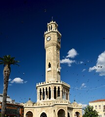 Fototapeta premium Historic Izmir Clock Tower with Pigeons in Flight Against Deep Blue Sky, Konak Square, Turkey