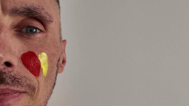 Close-up of a male fan of the national team applying make-up in the colours of the Spanish flag on his cheek before an important football match