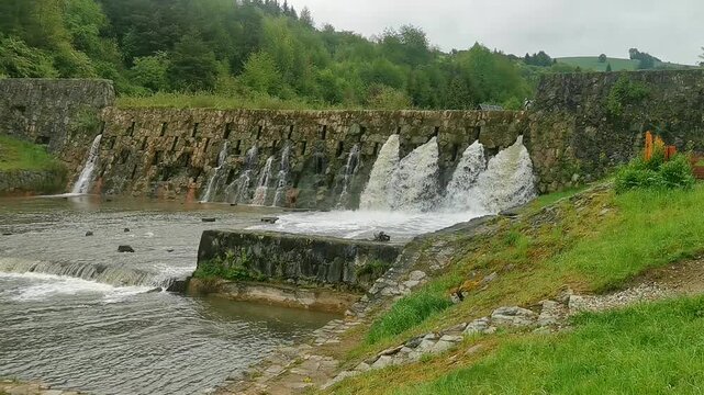 Water flowing over a stone weir on a mountain river, multiple streams cascading down the rustic dam in a rural landscape.