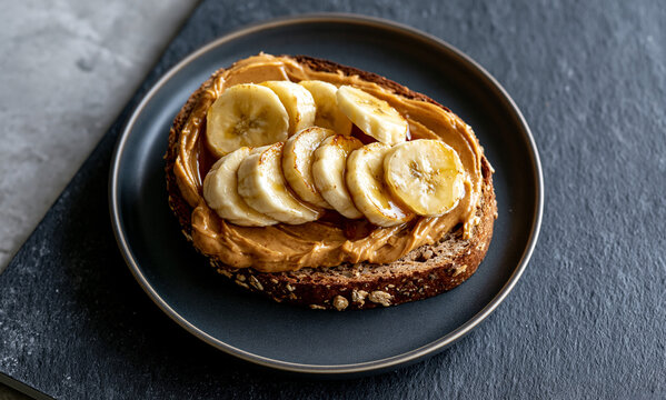 slice of dark toasted multigrain bread on a small dark ceramic plate on dark slate, spread with thick smooth golden-brown almond butter, topped with neatly arranged banana coins with slight caramelize