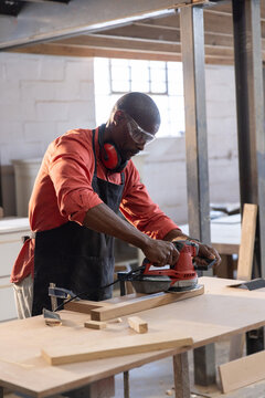 Red power sander smoothing long wooden plank on worktable inside workshop, with wood offcuts