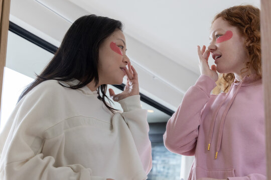 Two female friends sharing skincare in bright bathroom, wearing hoodies and pink heart eye patches