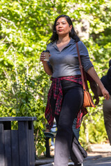 Fototapeta premium Adult female walking along tree-lined park path, holding paper coffee cup, wearing red plaid shirt
