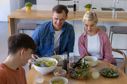 Parents with teenage son sharing pasta and salad at round wooden dining table in modern kitchen