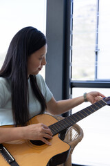 Fototapeta premium Mature African woman tuning wood cutaway guitar, sitting on chair by window, wearing watch
