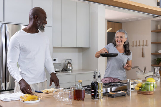 Diverse couple cooking in home kitchen, flipping pancake with frying pan and cutting bananas