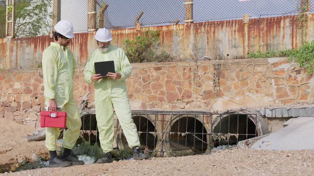 Two environmental scientists in protective gear inspect a sewer outfall, analyzing water quality and pollution data with a laptop for compliance.