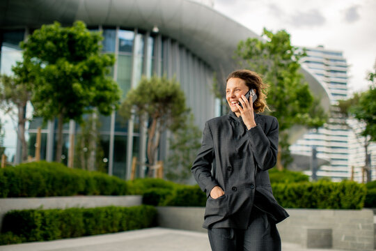 Business person talks on phone while walking in city near modern building and trees