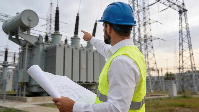Electrician wearing safety helmet and reflective vest holding project blueprint at power substation.