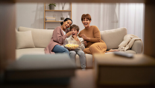 Same-sex parents and their son erupting into unfiltered, hysterical laughter at a chaotic comedy movie scene while playfully sharing a bowl of popcorn.