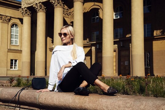 Blonde woman in sunglasses, white shirt, and black trousers sitting on a stone ledge in front of a grand neoclassical building with large columns