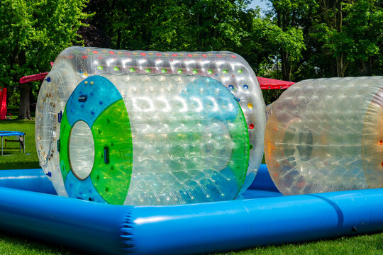 Two large, transparent inflatable water roller zorbing balls floating inside a blue inflatable pool on a green grass lawn surrounded by lush green trees on a bright sunny day.
