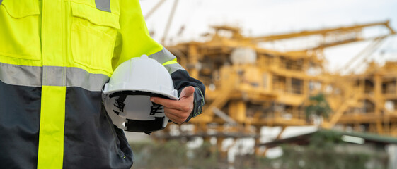 Fototapeta premium Industrial engineer in a high-visibility safety jacket and earmuffs, holding a hard hat and walkie-talkie radio at an offshore energy platform facility. Concept of workplace safety