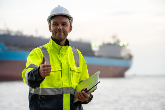 marine engineer or port worker wearing a hard hat and high-visibility jacket giving a thumbs up with a blurred container ship in the background. Maritime logistics safety and harbor operations 