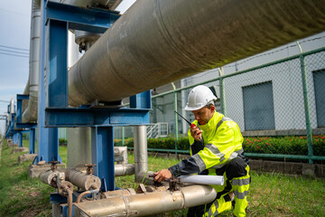 Naklejka premium industrial engineer wearing a white hard hat and high-visibility PPE squats to point at a large metal pipeline while speaking into a walkie-talkie during an outdoor refinery inspection.