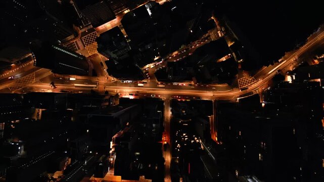 Aerial night artery of illuminated crossroad with golden traffic streams weaving between highrise blocks with neon reflections on asphalt. Dense urban fabric and metropolitan rhythm filmed from above
