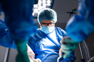 Naklejka na ściany i meble Focused senior male surgeon wearing glasses, blue scrubs, surgical mask and cap performs operation under bright LED light in hospital operating room.