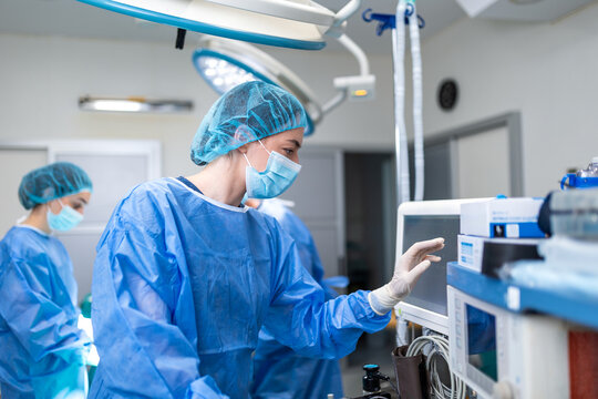 Professional female anesthesiologist in blue scrubs, mask, and sterile gloves adjusting medical equipment and monitoring vitals on a screen during a surgical operation in a hospital theater.