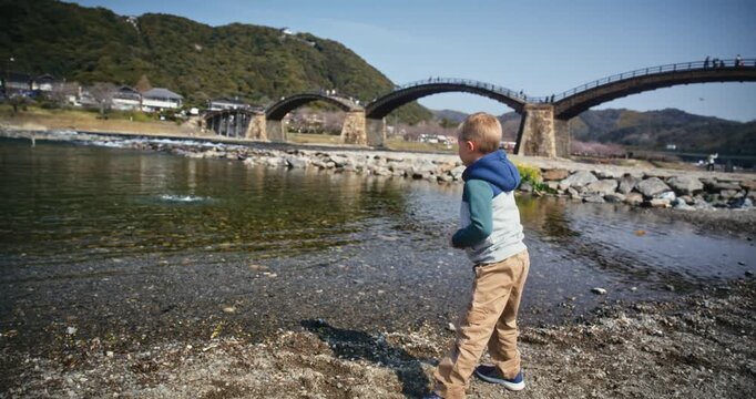 Young Boy Throwing Rocks at River Shore Near Arched Wooden Bridge In Japan