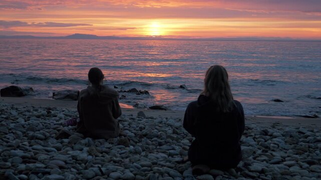Static handheld shot of two women on a pebble beach watching a stunning sunset over Lake Baikal. Vibrant pink sky reflects on rippling water with distant mountains.