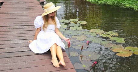 Fototapeten Lotusblume Woman sitting on wooden bridge by pond touching pink lotus flower, wearing white dress and straw hat, peaceful natural lighting, tropical vacation lifestyle, wellness and serenity.  © TravelMedia