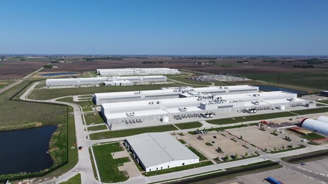 Forward drone shot above a large hyperscale data center campus moving toward a prominent power substation surrounded by agricultural land.