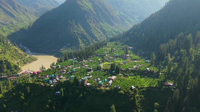 Riverside Himalayan village surrounded by orchards, dense conifer forests, and steep mountain slopes in the Parvati Valley near Kasol, Himachal Pradesh, India. Aerial drone footage, establishing shot