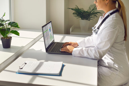 Doctor female uses laptop in office online. Medical professional types patient data on a computer, stethoscope nearby, desk in a bright workspace. Theme of telemedicine and digital workflow.