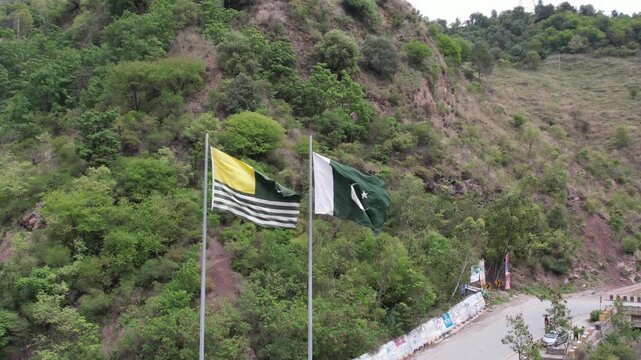 AJK and Pakistan flags fly beside a mountain road surrounded by green hillside vegetation, Pakistan
