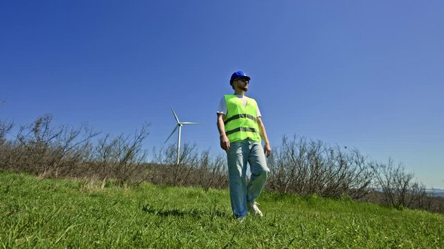 A windmill engineer walks through a green field on a sunny day. Wind turbines can be seen in the background. The engineer wears a safety vest and cap while inspecting the area.