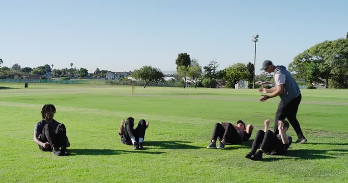Coach issuing signal and male trainees performing sit-ups for fitness on field, cricket stumps