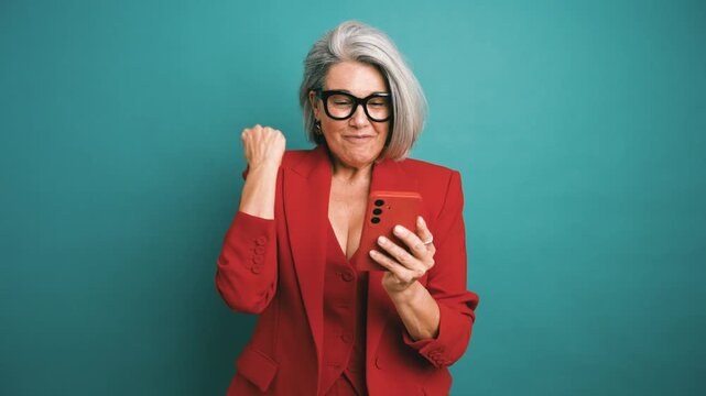 Elegant senior woman with grey hair receiving amazing news on her smartphone. She clenches her fist in victory, expressing pure joy and relief