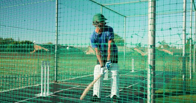 Anticipating bowl, male cricket player adjusting gloves, swinging bat in net cage, exiting field