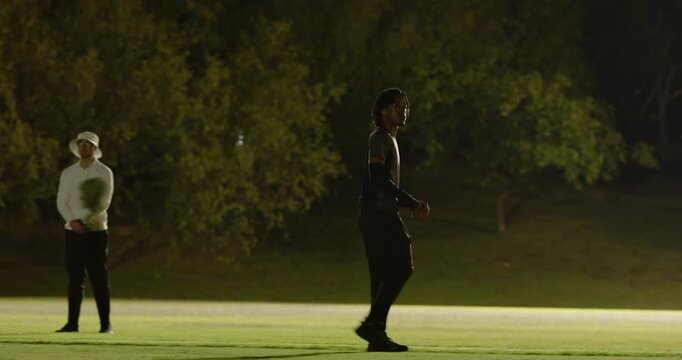 African american man grabbing ball tossing to friend practicing tosses, surveying floodlit field