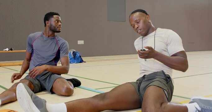 Catching breath after gym, african american friends scrolling device then stretching hamstring