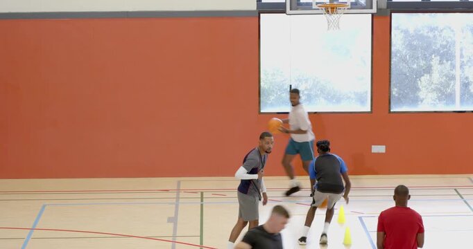 African american coach blowing whistle, guiding male players weaving cones making layups in gym